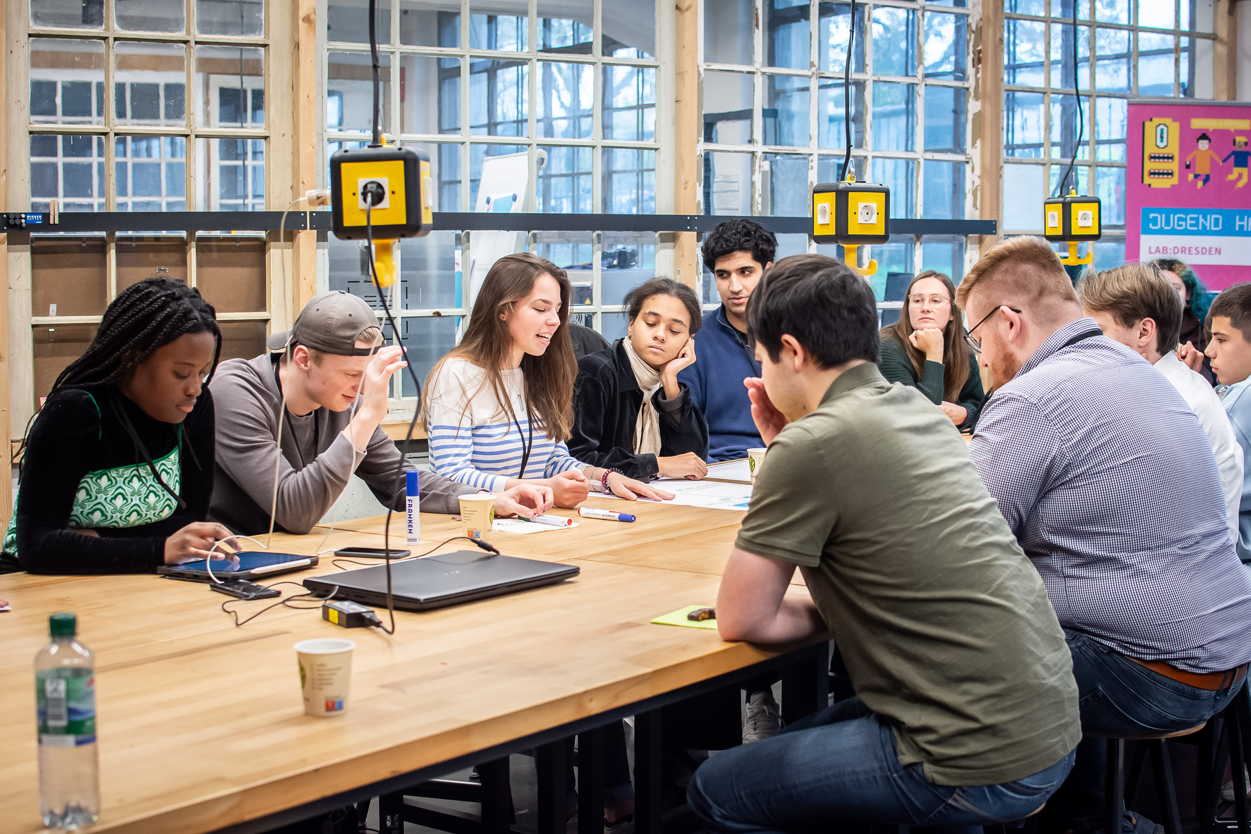 Auf dem Foto sieht man mehrere junge Menschen, die an einem großen Tisch in einem Büro sitzen und etwas besprechen.